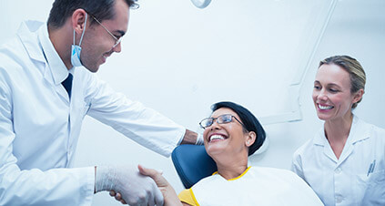 Female patient sitting in a dental chair being greeted by a male dentist with a female dental technician sitting next to her